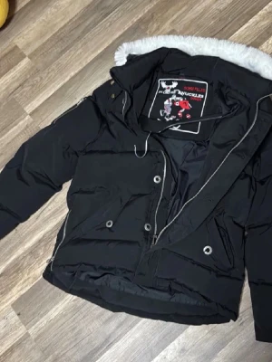 Svart Moose Knuckles dunjacka - Säljer en svart dunjacka från Moose Knuckles med vit fuskpäls på huvan och snygga metalldetaljer på ärmen. Jackan har dragkedja, knappar och flera fickor. Insidan har ett broderat hockeymotiv och tydlig logga. Perfekt för kalla dagar och riktigt stilren. Jackan är äkta också 