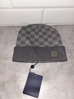 Grå rutig mössa från Louis Vuitton - Snygg grå mössa från Louis Vuitton med klassiskt rutigt mönster och ribbad uppvikt kant. Mössan har en metallbricka med Louis Vuitton Paris framtill och är tillverkad i ett mjukt, stickat material. Perfekt accessoar för att lyfta din vinterstil.