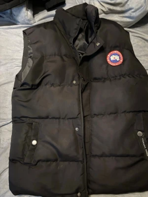 Svart dunväst från Canada Goose S - Svart dunväst från Canada Goose med klassisk logga på bröstet. Västen har hög krage, två fickor med tryckknappar och dragkedja framtill. Perfekt för lager-på-lager och håller dig varm under kyliga dagar.