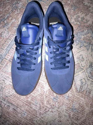 Adidas Gazelle marinblå sneakers - Säljer ett par Adidas Gazelle sneakers i marinblå mocka med ljusblå ränder och brun sula. Klassisk låg modell med snörning och rund tå. Perfekta för dig som gillar retrovibbar och stilrena detaljer.