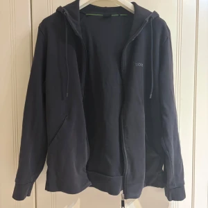 Mörkblå hoodie från BOSS - Snygg mörkblå hoodie från BOSS med dragkedja och huva. Tröjan har långa ärmar, snörning vid huvan och diskret BOSS-logga på bröstet. Tillverkad i mjuk bomull, perfekt för chill dagar. Stilsäker och enkel att matcha med jeans eller joggers. Priset kan diskuteras vid snabb affär