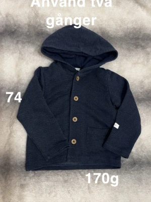 Mörkblå hoodie med knappar och huva - Mysig mörkblå hoodie för barn med träknappar framtill och en stor huva. Tröjan har en enkel ficka på ena sidan med diskret mönster. Perfekt för kyliga dagar och enkel att matcha.🌸Använd två gånger. Storlek 74