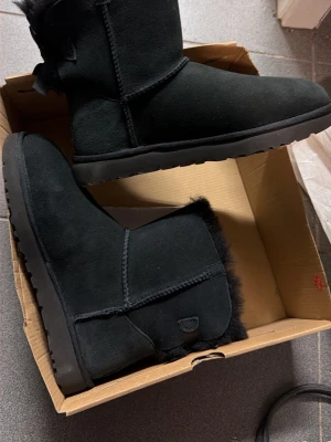 Svarta UGG boots med rosett - Helt nya, mamma köpte de men kom aldrig till användning, orginalpris 2099kr mitt pris 1599kr✅ Svarta UGG boots i mocka med fluffig insida och dekorativ rosett baktill. Klassisk rund tå och platt sula för skön komfort. Perfekta för kalla dagar och ger en mysig vibe till din outfit.
