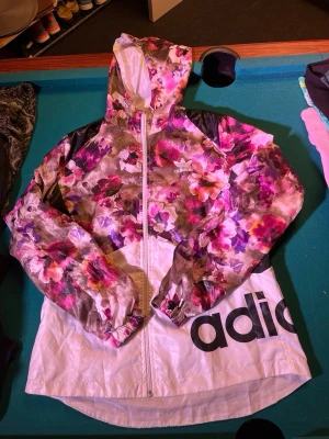 Blommig vindjacka från Adidas, strl L - Säljer en snygg vindjacka från Adidas i storlek L. Jackan har blommigt mönster i rosa, lila och beige toner, med vit nederdel och stor Adidas-logga. Utrustad med huva, dragkedja och elastiska muddar. Perfekt för dig som vill sticka ut.