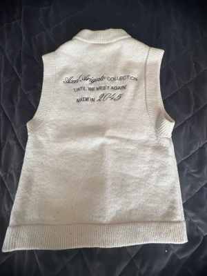 Vit stickad väst Axel Arigato - Säljer en clean vit stickad väst sense Axel Arigato med broderad logga på bröst. Västen har ribbade kanter vid hals, ärmhål och nederkantsöm. På ryggen finns cool tapetbro sil med texten 'Until alm, meet again'. Perfekt layering piece för dig som vill ha en stilren look.