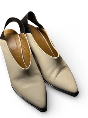Beige och svarta klackskor i skinn - Unika klackskor i beige skinn med svart bred rem över hälen och spetsig tå. Skorna har en hög klack och stilren design som sticker ut. Perfekta för dig som gillar minimalistiska men ändå edgy detaljer.