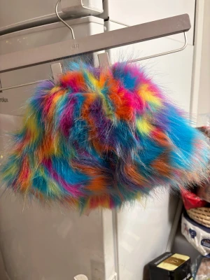 Färgglad fluffig hatt från  - Säljer en supercool hatt iregnbågens alla färger.Hatten är fluffig och har en riktigt statement look med blått, rosa, orange, gult och grönt. Perfekt för dig som vill sticka ut och älskar färgexplosioner!