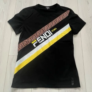 Svart Fendi t-shirt  - Säljer nu denna riktigt feta svarta t-shirt från Fendi med diagonal logga och färgglada ränder i vitt och gult. Brunt band med Fendi-monogram över bröstet. Äkta och köpt från Fendi. Den är knappt använd (max 2 gånger), storlek L otroligt mycket bra skick! Klassisk rund hals och korta ärmar. Tillverkad i mjuk bomull för skön känsla. Vid intresse bara att höra av sig! 