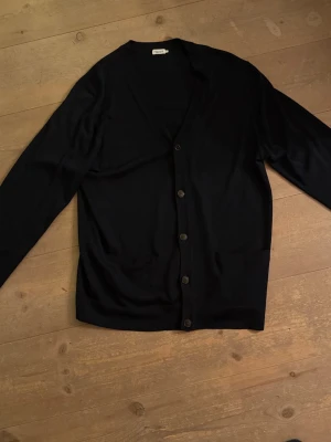 Marinblå kofta från Filippa K - Klassisk svart kofta från Filippa K med v-ringning och knäppning framtill. Tillverkad i mjuk bomull, har långa ärmar och två fickor framtill. Perfekt för lager på lager och enkel att styla till olika looks.