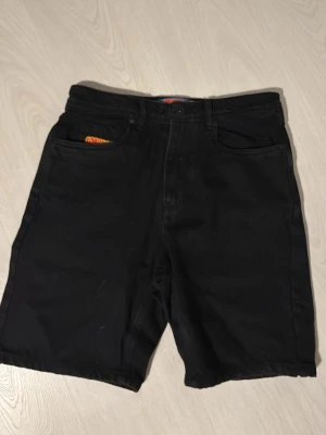 Svarta Empyre jorts  - Svarta shorts från GMPJRG med coola broderade loggor i rött och gult på fickan och bak vid midjan. Klassisk femficksdesign och knappstängning. Materialet är bomull och passformen är normal, perfekt för chill dagar.