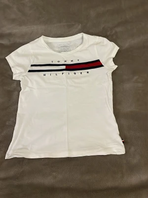 Vit Tommy Hilfiger t-shirt - Säljer denna Tommy Hilfiger tröjan, använd cirka 5 ggr, inga synliga täcken förutom storleken har tvättats bort, pris kan diskuteras