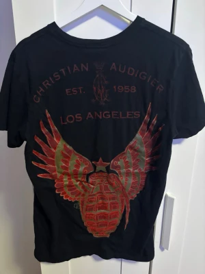 Christian Audigier svart t-shirt M - Svart t-shirt från Christian Audigier med stort tryck i rött och guld på både fram- och baksida. Framsidan har ett coolt skript-tryck och baksidan har en granat med vingar och texten 'Los Angeles'. Klassisk rund hals och korta ärmar.