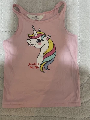 Rosa linne med enhörning från H&M - Säljer ett gulligt rosa linne från H&M med tryck av en enhörning i pastellfärger och texten 'positive mind' framtill. Linnet har smala axelband och är perfekt för varma dagar. Materialet är mjukt och skönt bomull.