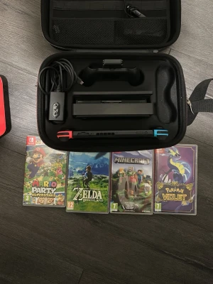 Nintendo Switch med andra tillbehör  - Nintendo Switch med blå och röd Joy-Con, originalförpackning, laddare, dockningsstation och bärväska. Fyra populära spel ingår: Mario Party Superstars, Zelda: Breath of the Wild, Minecraft och Pokémon Violet. Konsolen och tillbehören ser ut att vara i gott skick utan synligt slitage.