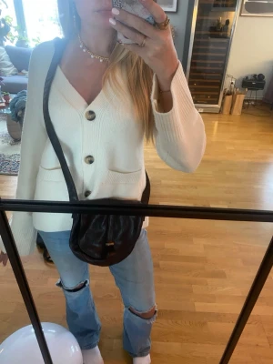 Brun axelväska från Complement - Säljer en brun axelväska från Complement i skinnimitation med rundad form och lock med magnetstängning. Väskan har ett justerbart axelband och en enkel detalj i metall framtill. Perfekt storlek för mobil, plånbok och nycklar.