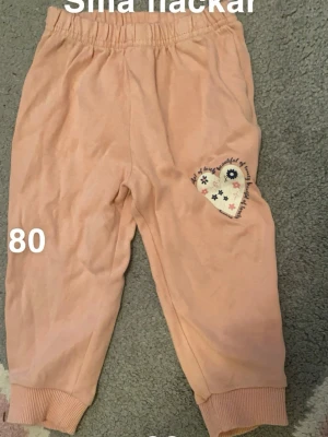 Byxor - Säljer ett par söta ljusrosa mjukisbyxor med resår i midjan och muddar vid bensluten. Byxorna har en hjärtformad detalj med blommigt mönster på ena benet. Perfekta för chill dagar hemma eller lek i parken.🌸Små fläckar. Storlek 80