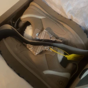 Beige sneakers från UGG med päls - Säljer ett par snygga beige sneakers från UGG med grov platt sula och fluffig vit päls på insidan. Skorna har meshdetaljer, mocka och coola mönstrade snören. Perfekt för dig som vill ha både stil och komfort.