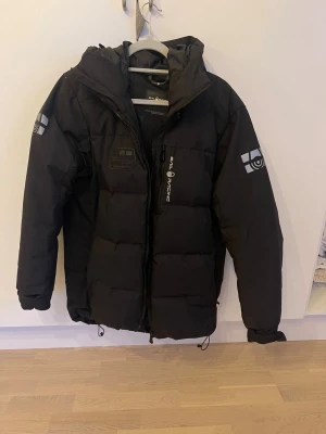 Svart dunjacka Sail Racing M - Svart dunjacka från Sail Racing med huva och dragkedja framtill. Jackan har coola loggor på ärmarna och bröstet, samt snörning nedtill för att justera passformen. Fylld med Allied Feather + Down, 500 fill power. Perfekt för kalla dagar. Jätte bra skick. Knappt använd