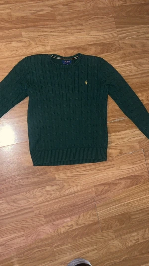 Grön kabelstickad POLO Ralph Lauren-tröja  - Supersnygg kabelstickad tröja från POLO Ralph Lauren i mörkgrönt. Klassisk modell med gul broderad logga. Tröjan är i väldigt fint skick, använd få gånger och utan defekter.