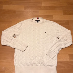 Vit kabelstickad tröja Tommy Hilfiger - Snygg vit kabelstickad tröja från Tommy Hilfiger med klassisk rund hals och diskret logga på bröstet. Tröjan har ribbade muddar och är långärmad, perfekt för dig som gillar stilrena och tidlösa plagg. Passar dig som vill ha en clean look med lite preppy vibe.