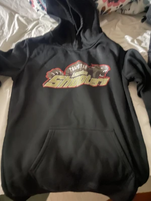 Trapstar London Shooters svart hoodie - Svart hoodie från Trapstar med stort London Shooters-tryck i gult, rött och grått på bröstet. Klassisk huva och magficka framtill. Perfekt för dig som gillar streetwear och vill sticka ut med ett coolt motiv.