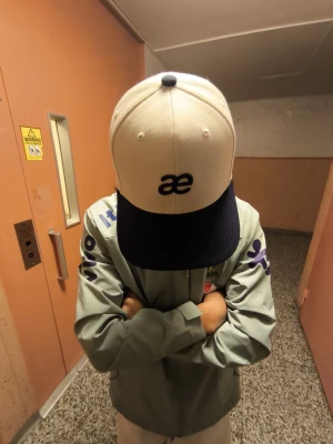 Beige och marinblå keps med AE-logga - Säljer en stilren keps i beige med marinblå skärm och broderad AE-logga framtill. Kepsen har klassisk form med justerbar rem bak och ventilerande hål. Perfekt för dig som vill ha en clean look med lite edge.