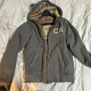 Grå hoodiejacka från Hollister - Säljer en grå hoodiejacka från Hollister med dragkedja och broderad CA-logga på bröstet. Jackan har ribbade muddar, två fickor fram och en huva med rutigt foder i beige och orange toner. Perfekt för chill dagar och streetwear.