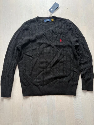 Svart kabelstickad tröja Polo Ralph Lauren - Svart kabelstickad tröja från Polo Ralph Lauren med klassisk rund hals och röd broderad logga på bröstet. Tröjan har ribbade muddar och är tillverkad i mjuk ull, perfekt för att hålla värmen under kyliga dagar.
