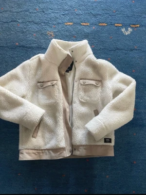 Vit teddyjacka från Vans XS - Säljer en vit teddyjacka från Vans i storlek XS. Jackan har hög krage, två bröstfickor med lock och knappar, samt sidofickor med beige detaljer. Mjuk och fluffig yta i fleece, perfekt för kyliga dagar. Snygg streetstil med Vans-logga på nederkanten. Jackan passar mig nästan som är 180cm lång och är S NYPRIS: 1249kr