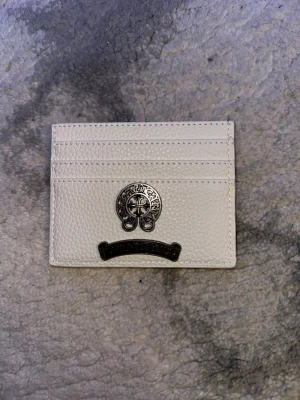 Vit plånbok från Chrome Hearts - Vit plånbok i skinn från Chrome Hearts med tre kortfack och snygga metall-detaljer framtill. Plånboken har en stilren och modern look med präglad yta och coola emblem i silver. Perfekt för dig som vill ha något unikt och lyxigt i fickan.