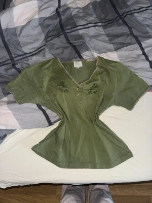 Olivgrön blus från Cressida, M - Olivgrön blus från Cressida med v-ringad hals och dekorativ spetskant i beige. Blusen har korta ärmar, tre knappar framtill och broderade blommor på bröstet. Tillverkad i mjuk bomull, perfekt för dig som gillar chill och söt stil.