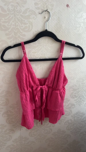 Rosa linnetopp med volanger och knyt - Säljer en söt rosa linnetopp med volanger och justerbara axelband. Toppen har en v-ringad front och knytning framtill för extra detalj. Materialet är lätt och krinklat, perfekt för varma dagar. Passar dig som gillar färg och en chill vibe.