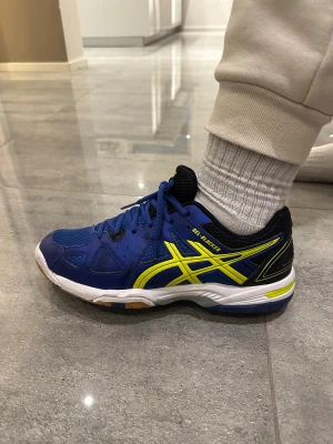 Blå ASICS Gel-Blocker sneakers - Säljer ett par blå ASICS Gel-Blocker sneakers med coola gula detaljer och svart mesh vid öppningen. Skorna är så gott som oanvända, super bra skick! Skorna har snörning, platt sula och slitstark gummisula med grymt grepp. Materialet är syntet och mesh, perfekt för dig som gillar sportig stil och komfort.
