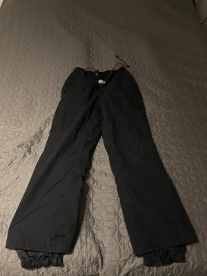 Svarta täckbyxor från Tuxer Woman - Svarta täckbyxor från Tuxer Woman med breda ben och elastisk midja med snörning. Byxorna har praktiska sidofickor med lock och dragkedja, samt resår vid bensluten för extra skydd mot snö och kyla. Perfekta för vinteraktiviteter.