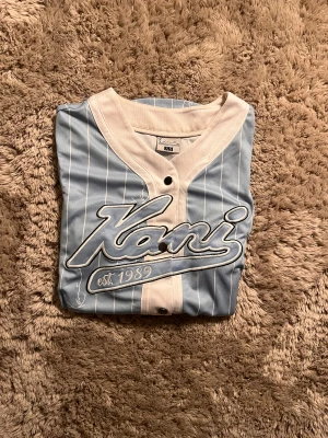 Blåvit randig skjorta från Karl Kani - Snygg baseball-inspirerad skjorta från Karl Kani i ljusblått med vita ränder och vit krage. Stort broderat Kani-tryck på bröstet och knappar framtill. Skjortan har korta ärmar och är gjord i mjukt syntetmaterial. Perfekt för dig som gillar streetwear och sportig stil.