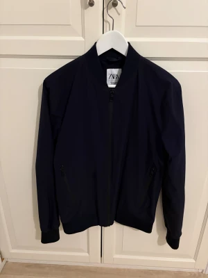 Svart bomberjacka från Zara - Snygg svart bomberjacka från Zara med klassisk rund krage och dragkedja framtill. Jackan har två fickor med dragkedja och ribbade muddar vid ärmslut och nederkant. Materialet är lätt och känns skönt mot kroppen, perfekt för dig som gillar stilren och enkel design.