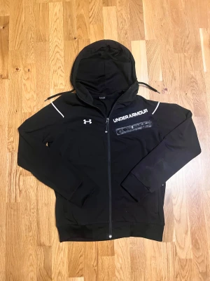 Svart vindjacka från Under Armour - Svart vindjacka från Under Armour med huva, vita detaljer och logga på bröstet. Jackan har dragkedja framtill, fickor med dragkedja och en sportig look. Perfekt för dig som gillar streetstyle och aktiva dagar. Snygg passform och stilren design.
