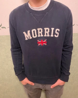 Mörkblå sweatshirt från Morris - Snygg mörkblå sweatshirt från Morris med broderad logga och brittisk flagga på bröstet. Tröjan har rund hals, långa ärmar och ribbade muddar. Perfekt för dig som gillar klassisk stil med en twist. Materialet är mjuk bomull som känns skönt mot huden.