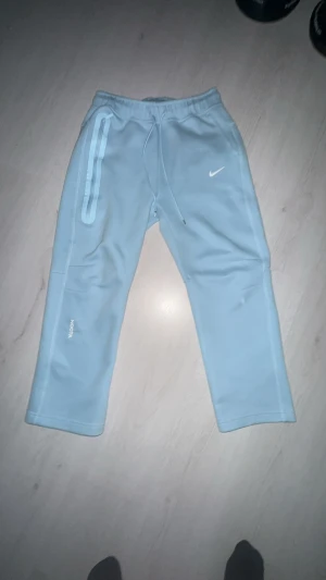 Nike NOCTA ljusblå mjukisbyxor M - Säljer ett par ljusblå mjukisbyxor från Nike NOCTA i storlek M. Byxorna har snörning i midjan, raka ben och coola detaljer med tryck på benen. Materialet är mjukt och skönt, perfekt för chill eller träning. Snyggt NOCTA-tryck och Nike-logga framtill.