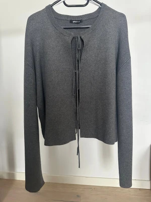 Grå kofta med snörning från Gina Tricot - Grå kofta från Gina Tricot i storlek L. Koftan har långa ärmar och snörning framtill istället för knappar. Materialet är stickat och känns mjukt mot huden.   Obs: Alla plagg säljs endast fram till 28/12