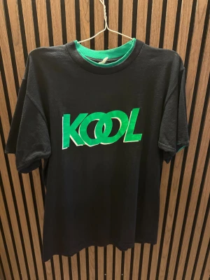 Svart KOOL t-shirt från Bluegrass - Svart t-shirt med grön och vit KOOL-print på bröstet från Bluegrass. Klassisk rund hals och korta ärmar. Materialet är mjuk bomull som känns skönt mot huden. Perfekt för dig som gillar retro och grafiska tryck.