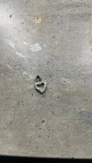 Hjärthänge i silver med glittriga stenar - Säljer ett sött hjärt berlock till halsband i silverfärg med glittriga stenar runt kanten. Perfekt att hänga på ett halsband för att ge din look lite extra bling. Hängsmycket har en öppen hjärtform och gnistrar snyggt i ljuset.