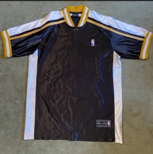 Champion NBA pikétröja svart/gul M - Snygg pikétröja från Champion med officiell NBA-logga. Tröjan är svart med vita och gula detaljer, har korta ärmar och ribbad krage med gula och vita ränder. Materialet är glansigt polyester och ger en sportig vibe. Perfekt för basketfans som vill sticka ut.