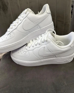 Nike Air Force 1 helvita sneakers - Nike Air Force 1 sneakers i helvit färg med klassisk låg siluett. Skorna har snörning, rund tå och platt sula med tydlig Air-dämpning. Tillverkade i skinn med perforerade detaljer på tån för extra ventilation. Perfekt för dig som gillar stilrena och tidlösa sneakers.