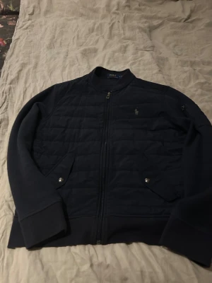 Mörkblå cardigan Jacka från Polo Ralph Lauren - Snygg mörkblå bomberjacka från Polo Ralph Lauren med quiltad front, ribbade muddar och dragkedja. Jackan har två fickor med tryckknappar och diskret logga på bröstet. Perfekt för dig som vill ha en stilren och klassisk look.