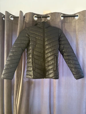 Svart pufferjacka från McKinley 158/164 - Säljer en svart pufferjacka från McKinley i storlek 158/164. Jackan har huva, dragkedja framtill och två fickor med dragkedja. Snyggt quiltad med reflekterande detaljer baktill. Perfekt för kyliga dagar och enkel att matcha med allt.