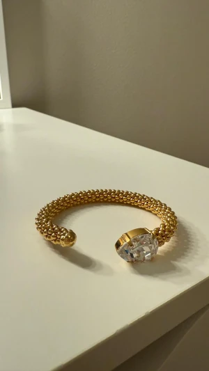 Guldfärgat armband Caroline Svedbom - Unikt armband från Caroline Svedbom i guldfärgad metall med pärlstruktur och en stor, gnistrande kristall i ena änden. Armbandet är öppet och har en lyxig känsla som verkligen sticker ut. Perfekt för dig som vill ha något extra till din look.