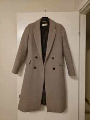 Beige lång kappa från MNG - Säljer en stilren beige kappa från MNG med dubbelknäppning och klassisk krage. Jackan är lång, har två fickor med lock och svarta knappar framtill. Perfekt för dig som gillar en clean och elegant look under höst och vinter.