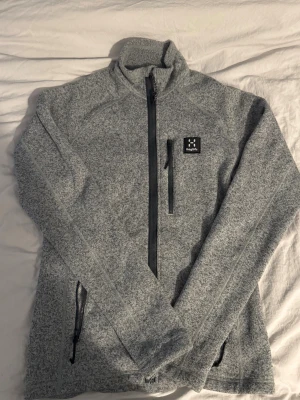 Grå fleecejacka från Haglöfs - Säljer en gråmelerad fleecejacka från Haglöfs med hög krage och dragkedja framtill. Jackan har tre fickor med dragkedja och en liten Haglöfs-logga på bröstet. Perfekt för kyliga dagar och outdoor-häng.
