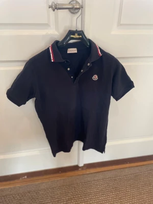 Blå pikétröja från Moncler - Tjena! Säljer en blå pikétröja från Moncler med klassisk krage och knappar framtill. Kragen har snygga rödvita ränder som ger en sportig vibe. Tröjan har korta ärmar och Moncler-logga på bröstet. Perfekt för dig som gillar stilrena märkesplagg. Liten skada vid knappen på första bilden (bild 4) Skick: 7/10. Storlek: xs. Bara att fråga om det är något! Priset går att diskutera!😊